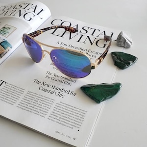 May include: Aviator sunglasses with a thin gold metal frame, a double bridge, and reflective blue lenses. The temples feature a brown and black tortoiseshell pattern. The sunglasses are positioned on an open magazine page with text.