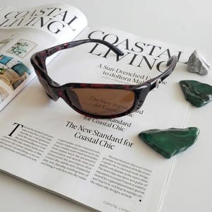 May include: Tortoiseshell-patterned sunglasses with dark brown lenses and a curved frame. The temples feature a small silver-toned metallic emblem near the hinges. The sunglasses are positioned on an open magazine page with printed text.