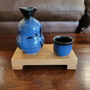 May include: A blue and black ceramic sake set with a small cup and a larger bottle, both with a black rim and a decorative design. The set is displayed on a wooden stand with a rectangular base.