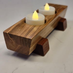 Puede incluir: Un portavelas de madera con dos velas LED. El soporte es de madera marrón clara y oscura, con forma rectangular y dos recortes circulares para las velas. Las velas tienen un brillo cálido y amarillo.