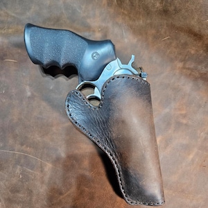 Op de afbeelding: Een bruine leren holster met een zilveren revolver erin. De holster heeft een gestikte rand en is ontworpen om strak om het pistool te passen.