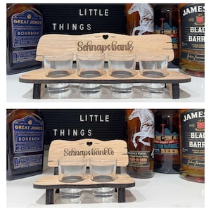 May include: A wooden shot glass holder shaped like a bench, with the text "Schnapsbank" engraved on the back. It holds four clear shot glasses on the top and four on the bottom. The background includes bottles of liquor and the words "Little Things".