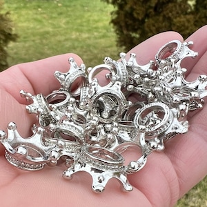 May include: A pile of silver crown-shaped charms, some with a raised design, others with a smooth surface. The charms are arranged in a hand, with some overlapping.