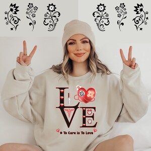 May include: A beige sweatshirt with a black "LOVE" graphic. The letters are decorated with red hearts, a stethoscope, a syringe, a bandage, a clock, and a medical symbol. The text "# To Care Is To Love" is printed below the graphic.
