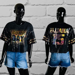 Black Sequin Top with Comp Day text design on the front in gold holographic spangle bling. The shirt back has custom text/name and numbers in gold holographic spangle bling. Gold bling stripes on sleeves all displayed on a black mannequin.