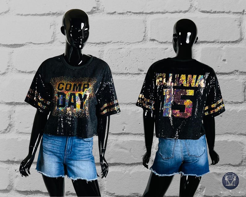 Black Sequin Top with Comp Day text design on the front in gold holographic spangle bling. The shirt back has custom text/name and numbers in gold holographic spangle bling. Gold bling stripes on sleeves all displayed on a black mannequin.