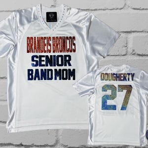 May include: Two white athletic jerseys. One jersey has "BRANDEIS BRONCOS SENIOR BAND MOM" in red and blue glitter lettering. The other jersey has "DOUGHERTY 27" in glitter lettering. Both jerseys have a V-neck.