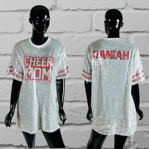 May include: A silver sequined t-shirt with the words "CHEER MOM" in red sequins on the front. The sleeves have red and white stripes. The back of the shirt has the name "RANIAH" in red sequins.