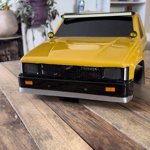 May include: Yellow toy truck with black details and a light bar on the roof. The truck has a black grill with the word "TOYOTA" printed on it. The truck is sitting on a wooden surface.