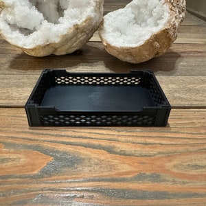 May include: A black plastic storage bin with a grid pattern on the sides. The bin is empty and sitting on a wooden surface.