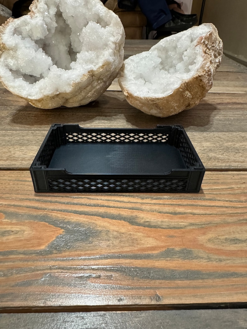 May include: A black plastic storage bin with a grid pattern on the sides. The bin is empty and sitting on a wooden surface.