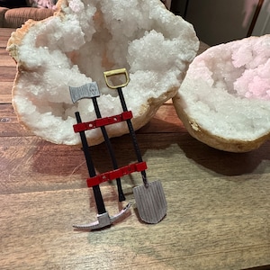 May include: Three miniature tools, a pickaxe, a shovel, and an axe, are mounted on a red wooden stand. The tools are made of metal and have a silver finish. The stand is attached to a large, white, crystal geode.