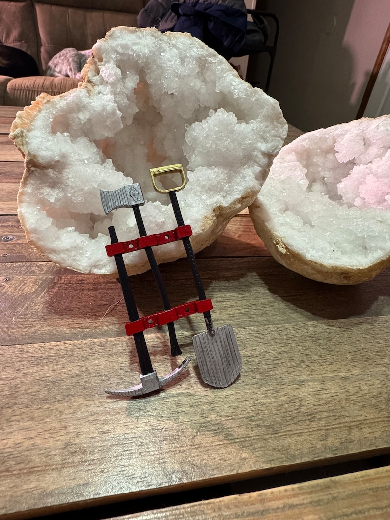 May include: Three miniature tools, a pickaxe, a shovel, and an axe, are mounted on a red wooden stand. The tools are made of metal and have a silver finish. The stand is attached to a large, white, crystal geode.