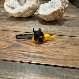 May include: A yellow toy chainsaw with a black handle and a silver blade. The chainsaw is lying on a wooden surface.