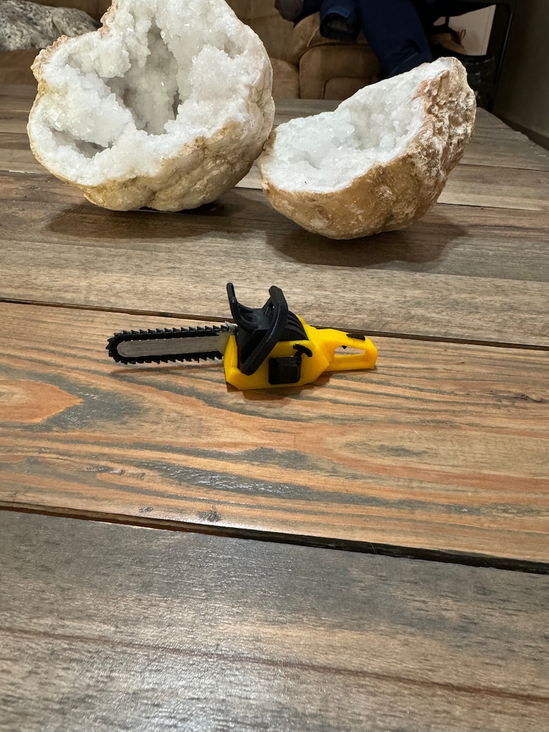 May include: A yellow toy chainsaw with a black handle and a silver blade. The chainsaw is lying on a wooden surface.