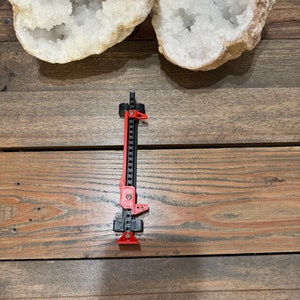 May include: A black and red jack stand with a metal chain and a red handle. The jack stand is on a wooden surface.