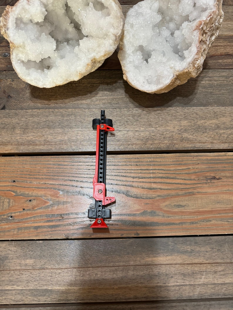 May include: A black and red jack stand with a metal chain and a red handle. The jack stand is on a wooden surface.
