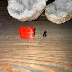 May include: A small, red plastic gas can with the word "GASOLINE" printed on it. The gas can is next to a small, black metal screw.