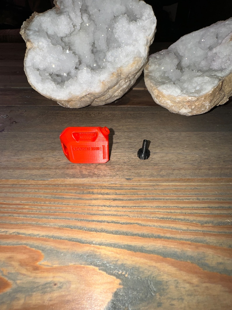 May include: A small, red plastic gas can with the word "GASOLINE" printed on it. The gas can is next to a small, black metal screw.