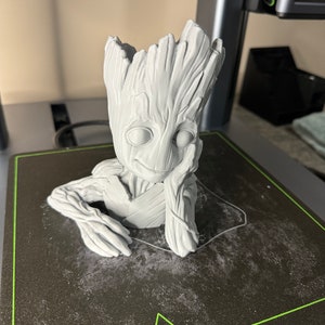 Puede incluir: Una figura gris impresa en 3D de un personaje de la franquicia de películas Guardianes de la Galaxia. El personaje está sentado con los brazos cruzados y tiene una cabeza grande con una textura de árbol. La figura está sentada sobre una superficie negra con el logotipo de AnkerMake.