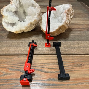May include: Three 3D printed jack stands, two red and one black, are arranged on a wooden surface. Two large, white, crystal geode halves are in the background.