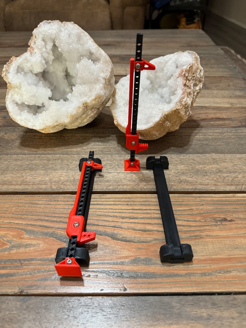 May include: Three 3D printed jack stands, two red and one black, are arranged on a wooden surface. Two large, white, crystal geode halves are in the background.