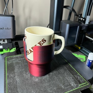 May include: A red 3D printed cup holder with a white ceramic mug inside. The mug has the text "Hayes" on it.