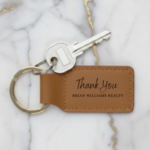 May include: A brown leather keychain with the words "Thank You" and "BRIAN WILLIAMS REALTY" printed in black. A silver key is attached to the keychain. The keychain is on a marble surface.