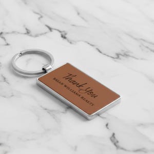 May include: A rectangular brown leather keychain with a silver metal frame and key ring. The text "Thank You" and "BRIAN WILLIAMS REALTY" are printed in black on the leather surface. The keychain is set against a marble background.