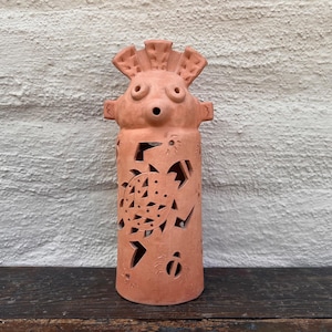 May include: A terracotta-colored ceramic sculpture with a cylindrical body and a stylized face. The sculpture features cut-out designs of a turtle and other shapes. The piece has a textured surface and is displayed on a wooden surface.