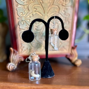 Puede incluir: Un par de pendientes con pequeñas botellas de vidrio con tapas de corcho. Cada botella contiene un pequeño objeto similar a un hueso. Los pendientes se muestran en un soporte de pendientes negro.