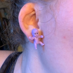May include: A pair of purple baby doll earrings. The dolls are wearing a white dress and have a pink face.