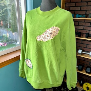 Puede incluir: Una sudadera de color verde lima con un parche de tela con estampado floral en la parte delantera. El parche tiene forma de nube.