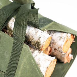 May include: A green canvas bag with straps and a buckle, filled with birch logs. The bag is designed for carrying firewood.