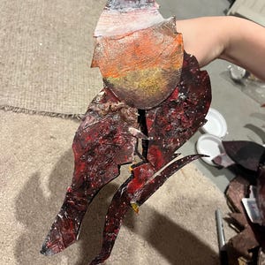 May include: A hand-painted, multi-layered cutout sculpture featuring a top section with blended white, orange, and yellow tones. The lower portion displays a dark, textured, and distressed surface with deep red, black, and brown paint splatters.
