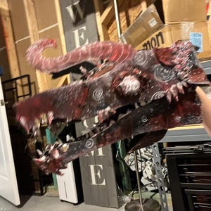 Può includere: Una maschera a testa di drago realizzata a mano con una superficie testurizzata in rosso scuro e nero. La maschera presenta corna ricurve prominenti, una mascella spalancata con denti affilati e intricati motivi a spirale impressi sull'esterno.