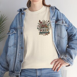 Puede incluir: Camiseta color crema con un diseño de cinta de cassette retro con el texto "Retro Echoes" y "Good Vibes". El diseño está rodeado de ilustraciones florales y de mariposas. La persona lleva una chaqueta vaquera azul claro.