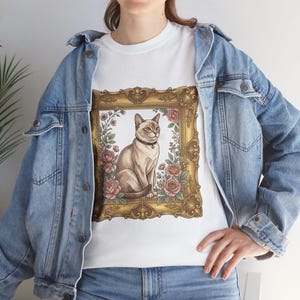 Puede incluir: Camiseta blanca con una ilustración enmarcada de un gato. El gato es de color marrón claro con un collar negro, enmarcado en un marco dorado adornado con rosas rosas y vegetación. La camiseta se usa con una chaqueta vaquera azul.