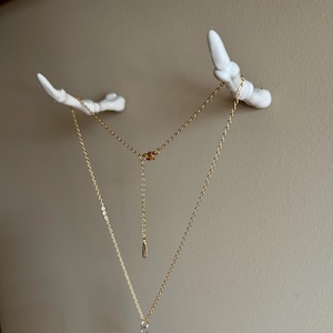 May include: A gold-colored metal chain necklace with a delicate link design, featuring an adjustable extender with a small metal tag. The necklace is draped across two white, matte-finished wall hooks shaped like skeletal finger bones.