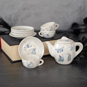 May include: A white ceramic tea set featuring a delicate blue rabbit and butterfly pattern with small gold dots. The set includes a teapot with a lid, multiple small cups, and matching saucers, all decorated with the same whimsical animal motif.