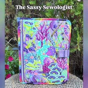 May include: A quilted fabric organizer titled The Sassy Sewologist and The Stitcher's Stash Book, featuring a vibrant purple background with a colorful underwater coral reef print, a vertical zipper on the left side, and a purple strap closure.
