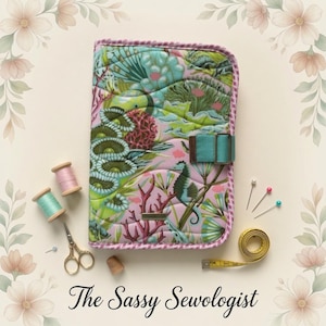 May include: A quilted fabric cover featuring a colorful coral reef print with seahorses, pink and green coral, and a teal strap closure. The cover is surrounded by two spools of thread, gold scissors, a thimble, pins, and a yellow measuring tape.