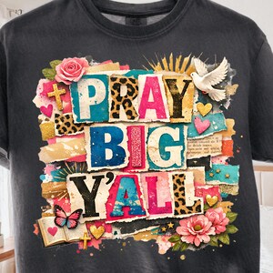 Könnte beinhalten: Dunkelgraues T-Shirt mit dem Schriftzug "PRAY BIG Y'ALL" in farbenfrohem, geschichtetem Text. Das Design umfasst florale und Leopardenmuster-Akzente, eine Taube, Herzen und einen Schmetterling. Das T-Shirt hängt an einem Holzkleiderbügel.