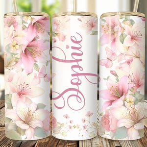 Op de afbeelding: Een bloemenbeker met roze lelies en vlinders op een witte achtergrond. De naam "Sophie" is in roze geschreven. De beker heeft een rietje en een goudkleurige rand.