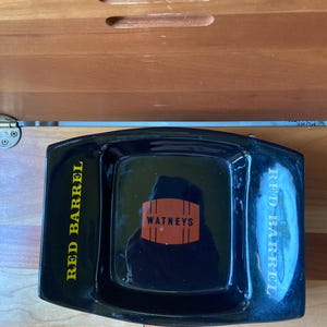 May include: A rectangular black ceramic ashtray featuring the text RED BARREL in yellow on the left side and white on the right side. The center of the ashtray displays an orange barrel graphic with the black text WATNEYS printed inside it.