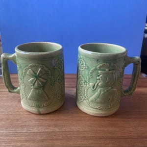 Könnte beinhalten: Zwei grüne Keramikbecher mit Henkeln. Ein Becher zeigt ein Windmühlendesign, der andere einen Mann, der eine Pfeife raucht. Beide Becher haben keltische Knotenmuster und stehen auf einer Holzoberfläche.