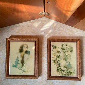 May include: Two rectangular wall plaques in brown wooden frames. The left plaque features a portrait of a person with curly hair wearing a light-colored garment. The right plaque depicts a person surrounded by green ivy vines and leaves.