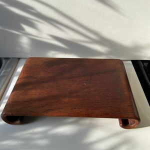 Puede incluir: Un soporte de madera marrón oscuro con bordes redondeados. El soporte está hecho de una sola pieza de madera y tiene una superficie lisa.