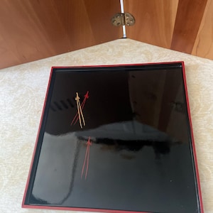 May include: A square black lacquer tray with a glossy finish and a thin red border. The surface features a hand-painted design of two crossed arrows in gold and red, along with a single vertical red arrow pointing upward toward the top edge.
