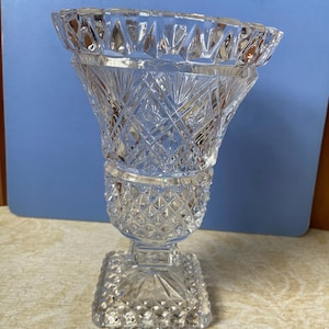May include: A clear, cut-glass vase with a square base and a flared top. The vase features intricate diamond and geometric patterns throughout its design. The vase is approximately 8 inches tall, perfect for displaying flowers or as a decorative piece.